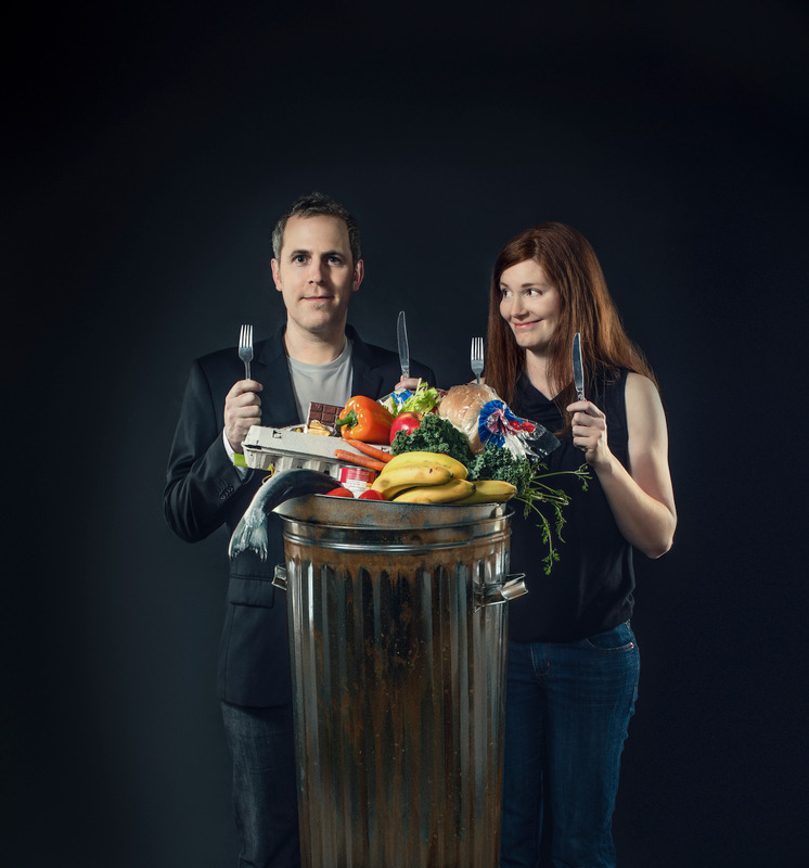 Photo of two people holding a knife and fork infront of a garbage can full of food